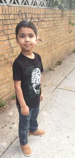 Young boy in urban outfit by a brick wall on a sunny day.