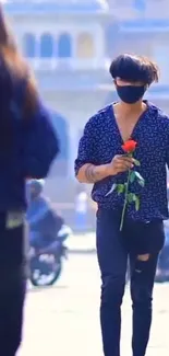 Masked man holding a red rose outdoors.