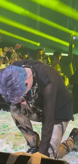 Blue-haired performer kneeling during a vibrant concert with neon lights.