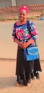 A woman smiling in a floral dress with a blue bag on a sunny street.