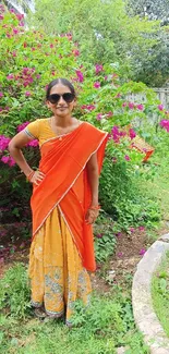 Woman in colorful saree standing in lush green garden with pink flowers.