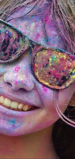 Smiling person covered in bright Holi colors with reflections in sunglasses.