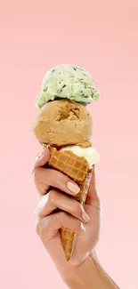 A hand holding a triple scoop ice cream cone against a pink background.