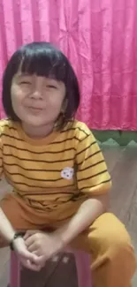 A smiling child in a striped shirt against a pink curtain backdrop.