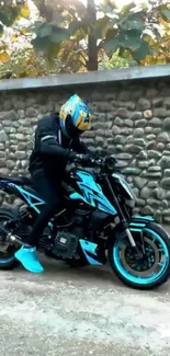 Motorcycle rider with turquoise bike near stone wall.