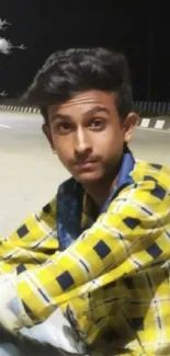 Young man in yellow plaid shirt sitting on a quiet night street.