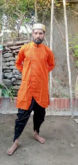 Man in orange ethnic attire stands in rustic yard.