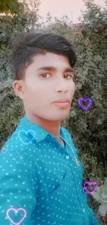 Young man poses outdoors in teal shirt, surrounded by greenery.