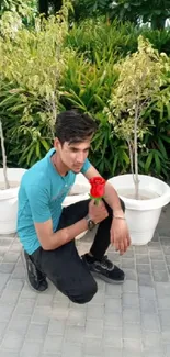 Man holding a red rose kneeling outdoors next to vibrant green plants and white pots.