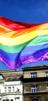 Vibrant rainbow flag flying high against a clear blue sky with buildings below.