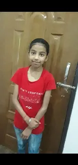 Girl in a red shirt posing in front of a brown wooden door.