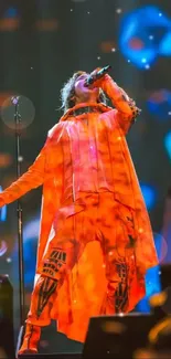 Performer in vibrant orange outfit singing energetically on stage with blue backdrop.