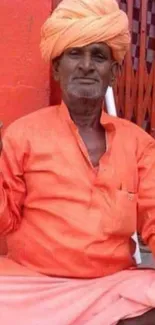 Man in vibrant orange traditional attire with orange turban seated.