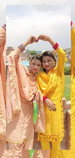 Two women in colorful traditional attire forming a heart shape with hands.