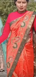 Person wearing a vibrant orange saree with intricate designs outdoors.