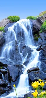 Vibrant waterfall with lush greenery and yellow flowers under a clear blue sky.