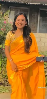 Smiling woman in a vibrant yellow saree holding a phone outdoors.