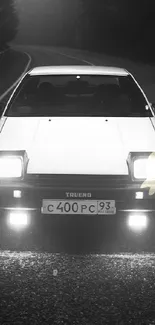 Vintage car with headlights on foggy road at night in grayscale.