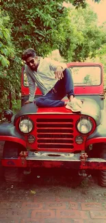 Man posing on red vintage Jeep in lush outdoor setting.