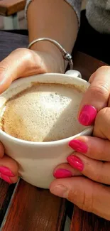 Hands with pink nails holding a coffee cup on a wooden table.