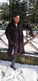 Man in dark coat stands in snowy forest setting with wooden fence and trees.