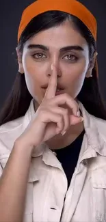 Woman with orange headband on a dark background, finger on lips.