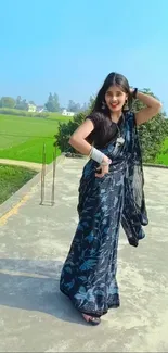 A woman in a vibrant saree poses outdoors against a rural backdrop.