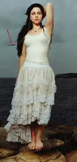 Woman in white dress on rocky shore with an ocean backdrop.