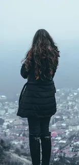 Woman in black coat overlooking a foggy cityscape from a high viewpoint.