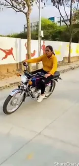 Woman rides motorcycle on urban street with colorful wall mural.