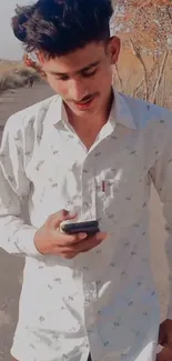 Young man with phone on sunlit rural road, wearing a white shirt.