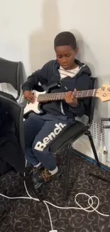 Young boy playing electric bass guitar intensely seated on a chair.